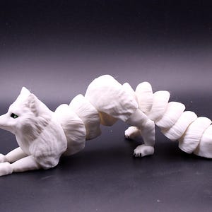 May include: A white sculpture of a fox with a segmented body, lying down with its head and front paws raised. The fox has green eyes and is set against a dark background. The sculpture is a unique art piece.