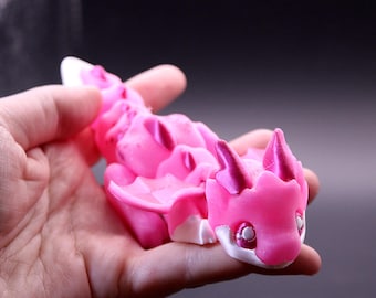May include: A pink and white articulated dragon figurine. The dragon has a detailed design with horns, wings, and a segmented body. The figurine is held in a hand, showcasing its size and intricate features. The dragon's eyes have a metallic sheen.