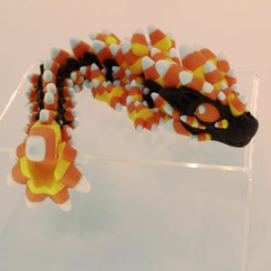 May include: A 3D printed articulated dragon figurine featuring a black base colour with a candy corn pattern of orange, yellow, and white triangular spikes covering its body. The dragon has large orange eyes and a segmented, flexible tail structure.