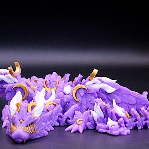 May include: A purple articulated dragon figurine featuring white wing accents and gold crescent moon symbols along its back and head. The creature has a textured, scale-like body with a detailed face, pointed ears, and a long, segmented tail.