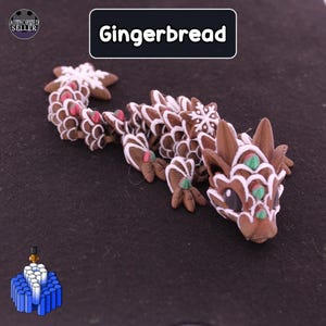 May include: A brown articulated dragon figurine with white icing-like patterns, small green and red accents, and a star-shaped tail tip. The text "Gingerbread" is displayed in a black box above the dragon, along with an "AUTHORIZED SELLER" logo.