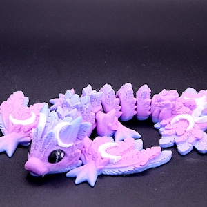 May include: A flexible, articulated dragon figurine in shades of pastel purple and blue. The creature features white crescent moon symbols on its head and wings, large black eyes, and a segmented body with textured, leaf-like scales throughout.