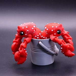 May include: Two red and white spotted toy crabs are holding onto the rim of a gray plastic bucket.