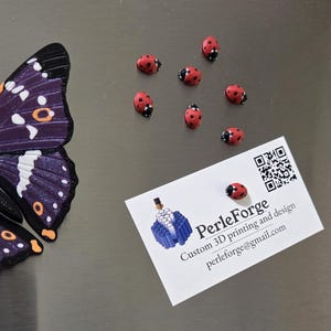 May include: A purple and black butterfly wing, seven small red ladybug figurines with black spots, and a white business card with a QR code, the text PerleForge, Custom 3D printing and design, and the email address perleforge@gmail.com.