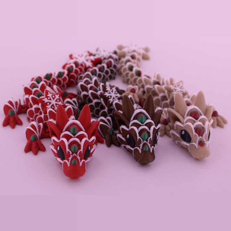 May include: Three articulated dragon figurines featuring a scale-like texture with white snowflake patterns. The dragons are colored in red, dark brown, and light tan, each with small green accents on the scales and large black eyes.