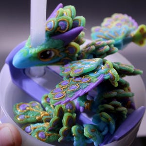May include: A detailed, colorful dragon figurine in shades of blue, green, purple, and gold. The dragon has intricate feather-like details and is perched on a white and purple base. The figurine is a fantasy-themed collectible.