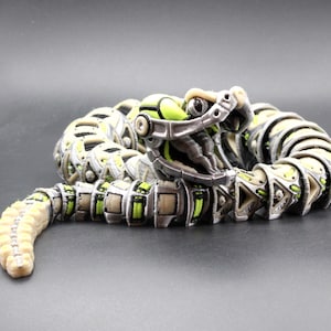 Könnte beinhalten: Eine detaillierte, bewegliche Schlangenfigur mit mechanischer Ästhetik. Die Schlange hat einen segmentierten Körper in Off-White, Grau und Grün mit aufwendigen Details. Der Kopf ist geöffnet und zeigt das mechanische Design.