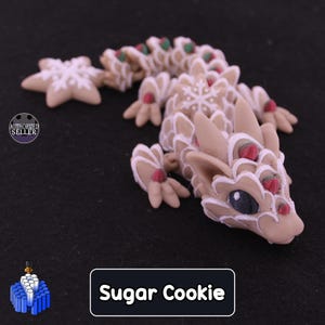 May include: A tan articulated dragon figurine with white icing-like piping, red and green accents, and a white snowflake pattern on its back. The tail ends in a star-shaped cookie. A label at the bottom reads Sugar Cookie.