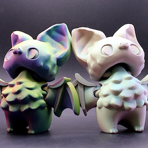May include: Two bat figurines. One is a gradient of green, purple, and blue. The other is off-white. Both have large ears, open mouths, and outstretched wings. The figurines are standing side-by-side.