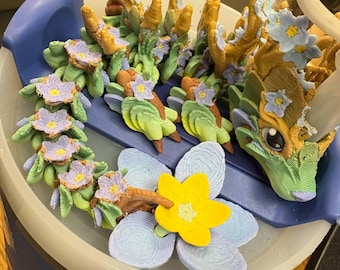 Può includere: Una scultura di drago stampata in 3D con un design floreale. Il drago presenta un corpo verde, corna marroni e fiori viola e gialli. La scultura è su una superficie blu e bianca.