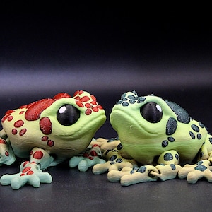 Peut inclure: Deux petites figurines de grenouilles imprimées en 3D. Une grenouille est verte avec des taches noires et l'autre est verte avec des taches rouges.