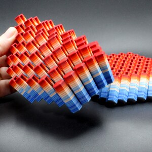 May include: A 3D-printed geometric sculpture composed of numerous small, rectangular blocks. The blocks are arranged in a layered pattern, with a gradient of colors from red to blue. The sculpture is held by a hand, and another similar piece is nearby.