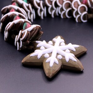 May include: A garland featuring brown, textured, gingerbread-style star shapes decorated with white icing patterns. Each star is connected to the next, with a prominent white snowflake design on the front of the lead star piece.