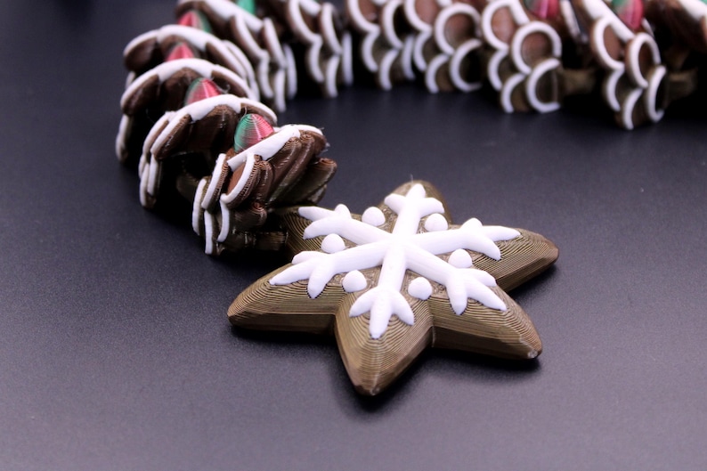 May include: A garland featuring brown, textured, gingerbread-style star shapes decorated with white icing patterns. Each star is connected to the next, with a prominent white snowflake design on the front of the lead star piece.