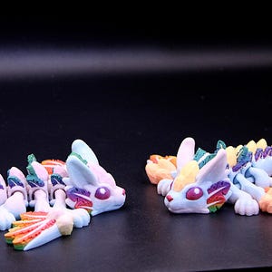 May include: Two colourful, segmented, articulated, dragon-like figures with rainbow-coloured scales and wings. The figures are made of a white material and have a whimsical, playful design.