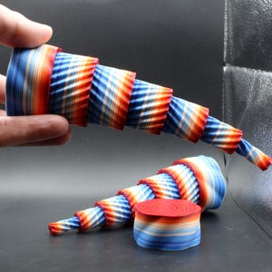 May include: A pair of abstract, spiral-shaped sculptures in red, white, and blue. One is held by a hand, the other rests on a surface with a small, circular stack of matching material. The designs feature layered, textured patterns.