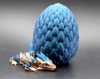 May include: A 3D printed articulated dragon figure with tan and blue scales resting beside a large, textured blue egg. The dragon has a detailed head with visible eyes and a segmented body, while the egg features a repeating scale pattern.