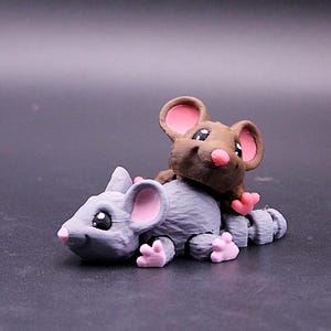 Puede incluir: Una figura impresa en 3D de dos ratones. Un ratón es gris con orejas y patas rosas, y el otro es marrón con orejas y patas rosas. El ratón marrón está posado sobre la espalda del ratón gris. La figura está sobre una superficie oscura.