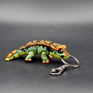 May include: A green, orange, and yellow articulated lizard keychain. The lizard has a segmented body, with orange and black detailing on its back. It has a silver keyring and clasp attached to its head.