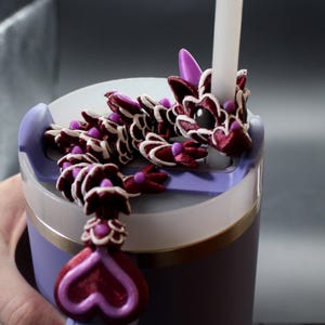 May include: A purple and white tumbler with a decorative dragon lid. The dragon is burgundy with white accents and purple details. A white straw is inserted through the dragon's head. The tumbler has a gold band near the bottom.
