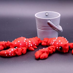 May include: Two red lobster figurines with white spots, lying on a black surface. A small gray bucket is in the background.