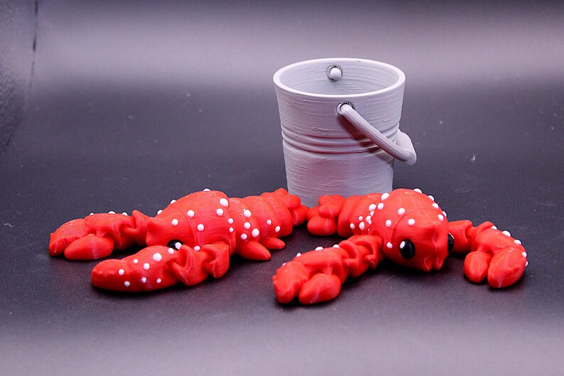 May include: Two red lobster figurines with white spots, lying on a black surface. A small gray bucket is in the background.