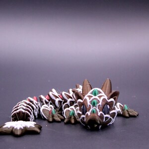 May include: A brown articulated dragon figurine featuring white painted edges on its scales, dark purple eyes, and small green accents on its head and body. The creature has a segmented tail and a detailed head with two pointed ears.