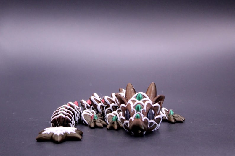 May include: A brown articulated dragon figurine featuring white painted edges on its scales, dark purple eyes, and small green accents on its head and body. The creature has a segmented tail and a detailed head with two pointed ears.