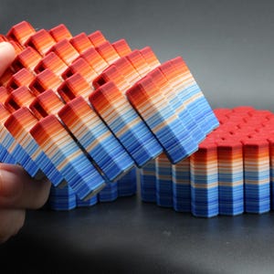 May include: A collection of colorful, layered geometric shapes in shades of red, orange, white, and blue. One set is held by a hand, while another sits on a dark surface. The layered design creates a textured, three-dimensional effect.