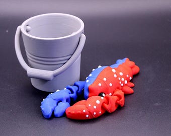 May include: A grey plastic bucket with a handle beside a red and blue lobster figurine. The lobster has white spots. The bucket and lobster are on a dark surface.