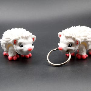 Op de afbeelding: Twee kleine witte egelbeeldjes met gestructureerde stekels, zwarte ogen en roze neuzen en pootjes. De ene egel is een losstaand figuur, terwijl de andere een zilveren metalen sleutelring in de mond heeft en fungeert als een sleutelhanger.