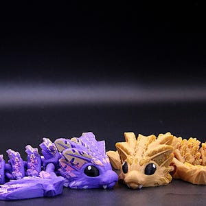May include: Two small, segmented dragon figurines. The dragon on the left is purple with pink accents. The dragon on the right is a light brown colour.