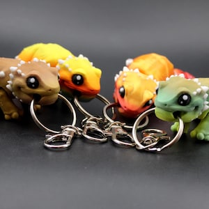 May include: A collection of colorful lizard keychains. Each keychain features a detailed, miniature lizard figurine in shades of brown, yellow, orange, and green, with white accents. The lizards have large, black eyes and are attached to silver key rings.