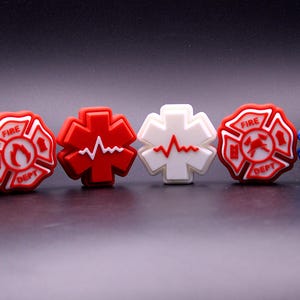 Puede incluir: Una colección de amuletos coloridos con temática de servicios de emergencia. Incluye un amuleto blanco con un gráfico del 911, amuletos rojos y blancos del departamento de bomberos, un amuleto de frecuencia cardíaca rojo y blanco y un amuleto con símbolo médico azul.