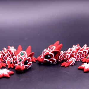 May include: Two articulated dragon figurines crafted in red with white scale detailing and dark eyes. Each dragon features a long, segmented body with small green accents and white snowflake-shaped ornaments attached to the sides and tail.