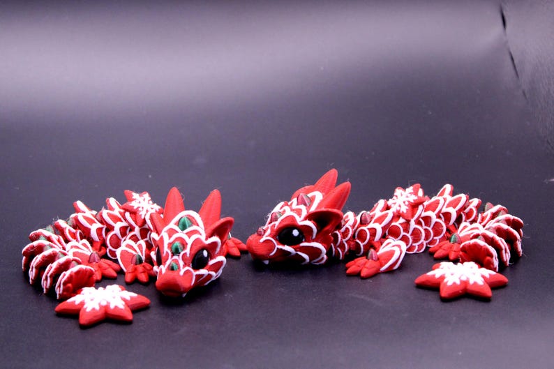May include: Two articulated dragon figurines crafted in red with white scale detailing and dark eyes. Each dragon features a long, segmented body with small green accents and white snowflake-shaped ornaments attached to the sides and tail.