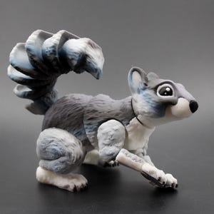 May include: A detailed, three-dimensional squirrel figurine in shades of gray, white, and blue. The squirrel has a large, fluffy tail and is posed in a dynamic, forward-facing stance. The figurine is likely made of a hard plastic or resin material.