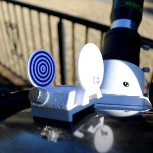 May include: A white telescope with a blue and white target sight. The telescope is mounted on a black surface.