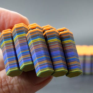 Puede incluir: Una mano sostiene varios objetos hexagonales multicolores. Cada objeto está compuesto por numerosas capas horizontales en tonos de azul, naranja, amarillo y verde. Los objetos tienen un aspecto texturizado y están apilados.