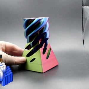 May include: A 3D printed object with a pyramid-shaped base featuring green and pink sides with oval cutouts. The top section is a spiraling, multi-colored structure transitioning through shades of blue, purple, and teal. A small graphic icon is visible.