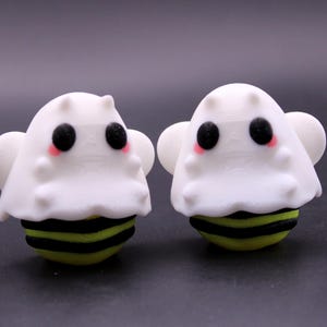 May include: Two novelty ghost-bee figurines. They have white bodies, black eyes, and rosy cheeks. The lower part of the bee is yellow with black stripes. The background is dark.