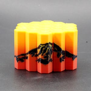 May include: A 3D-printed, geometric block with a sunset-themed design. The block features a gradient of colors, transitioning from red at the base to yellow at the top. A black mountain silhouette is printed on the front.