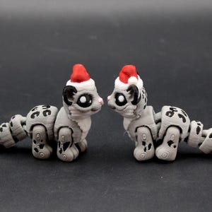 May include: Two articulated gray leopard figurines with black spots and red Santa hats. The figurines are facing each other, with a dark background. The leopard figurines are a unique Christmas decoration.
