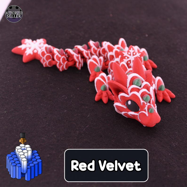 May include: A 3D printed articulated dragon figurine in red with white trim and green accents. The dragon features snowflake patterns on its back and tail. A black rectangular label with white text reads Red Velvet.