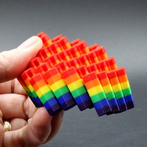 May include: A colorful, geometric sculpture in a rainbow of hues. The object is composed of stacked, rectangular blocks, each displaying a spectrum of colors from red to violet. The design is held in a hand against a gray background.