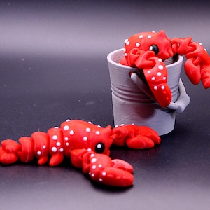 May include: Two red and white spotted lobster figurines. One lobster is sitting in a gray bucket, the other is lying on a black surface.