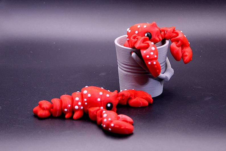 May include: Two red and white spotted lobster figurines. One lobster is sitting in a gray bucket, the other is lying on a black surface.