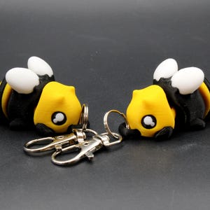 May include: Two bee-shaped keychains. Each keychain features a yellow head, black body with yellow stripes, white wings, and a silver keyring. The keychains are lying on a dark surface.