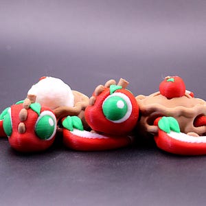 以下が含まれることがあります： 奇抜な、3Dプリントされた食品をテーマにしたフィギュアのコレクション。セットには、緑の目と葉を持つ赤いリンゴのような生き物、上に赤いリンゴが乗ったパイ、白いアイスクリームのスクープが含まれています。