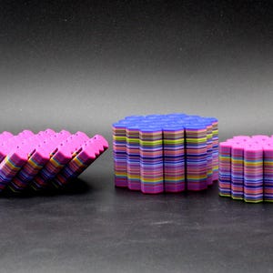 May include: Three stacks of colorful, geometric shapes. The shapes are in shades of pink, blue, yellow, and green. The stacks are arranged on a dark surface. The shapes appear to be made of layered material.